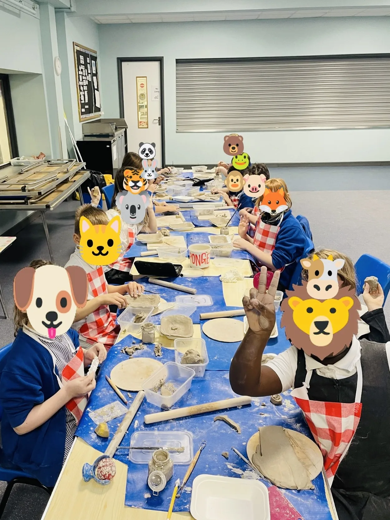Children in gingham aprons making clay pieces around a long table at the after-school clay club, faces covered with animal emojis