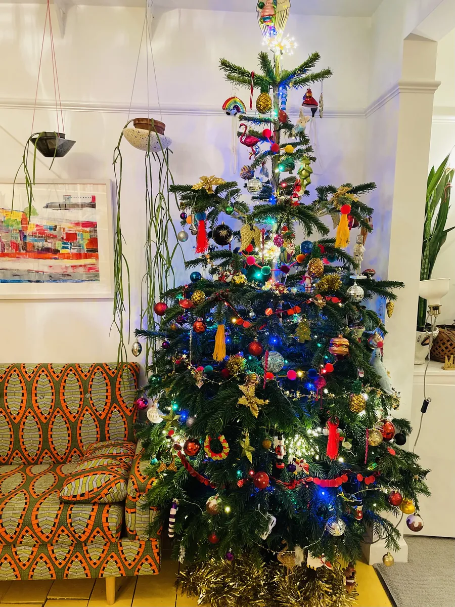 A Christmas tree full of bright baubles and pom-poms next to a print sofa