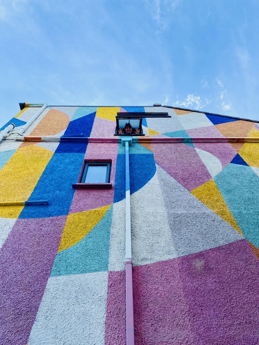 A house wall painted in pink, blue, yellow and teal shapes, with Gemma waving from the top window